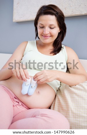 Close up portrait view of a young pregnant woman and mother to be sitting on her bed at home, holding her baby boy blue booties on her belly and smiling.