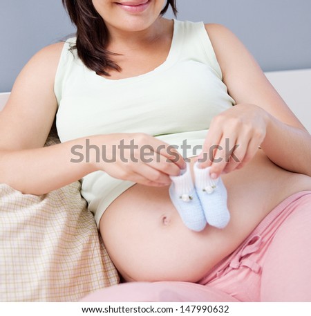 Close up view of a young pregnant woman and mother to be sitting on her bed at home, holding her baby boy blue booties on her belly and smiling.
