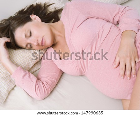 Over head view of a young pregnant woman and mum to be sleeping on a bed at home, relaxing and resting while wearing a pink top and white shorts, interior.