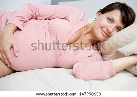 Close up portrait of a beautiful pregnant woman laying down on a bed at home, resting and relaxing and proudly smiling at the camera, interior.