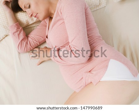 Over head profile view of a young pregnant woman and mum to be sleeping on a bed at home, relaxing and resting while wearing a pink top and white shorts, interior.