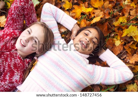 Two diverse teenage girls friends relaxing on a bed of autumn leaves in a park, smiling at camera and having fun during a day out in the fall season, outdoors.