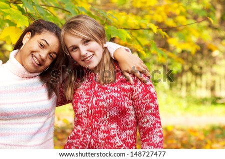 Portrait of two diverse teenager best friends with their arms around each other shoulders, in a beautiful park during fall season with orange, yellow and brown leaves all around them.