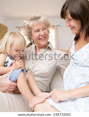 Grand mother, pregnant daughter and child grand daughter sitting together as a generations family on a white sofa at home and having a good time. Smiling and being happy indoors.