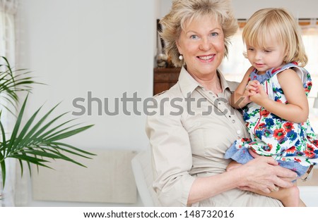 Attractive grand mother carrying her young grand daughter in her arms while standing in a living room at home, being proud and smiling at camera. Indoors.