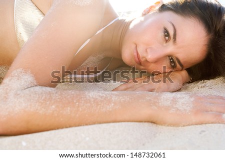 Close up portrait of a sexy young woman laying down on a white sand beach wearing a luxurious sequins golden bikini with the sun rays shining at sunset while on vacation.