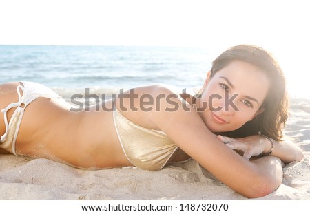 Side beauty portrait of a young attractive woman laying down and relaxing lounging on a white sand beach shore wearing a gold sequins bikini against the sunset, being sexy and smiling.
