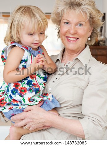 Attractive grand mother carrying her young grand daughter in her arms while standing in a living room at home, being proud and smiling at camera. Indoors.