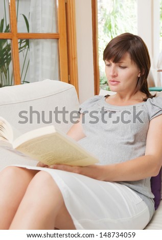 Attractive pregnant young woman sitting down and relaxing on a sofa at home while reading a book during a sunny day with garden views. Interior.
