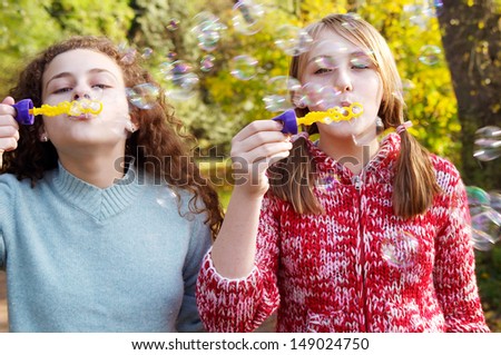 Frontal view of two teenager friends playing games together and blowing soap bubbles up in the air while in a natural park during a fall autumn sunny day, outdoors.