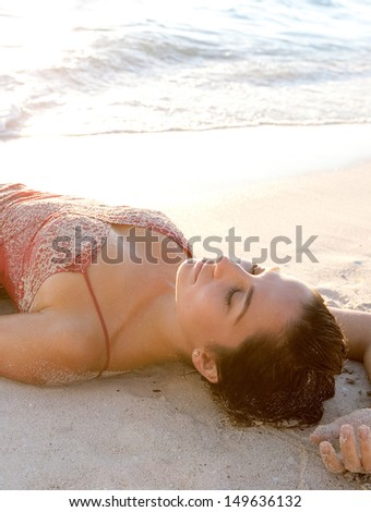 Side view of a beautiful woman laying down on the shore of a white sand beach with her arm up, bathing and relaxing while dressed in pink at sunset during a summer vacation.
