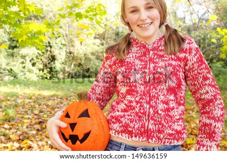 Joyful girl holding a fake halloween pumpkin under her arm and having fun while visiting a forest park with falling leaves and autumn colors during a sunny holiday day.
