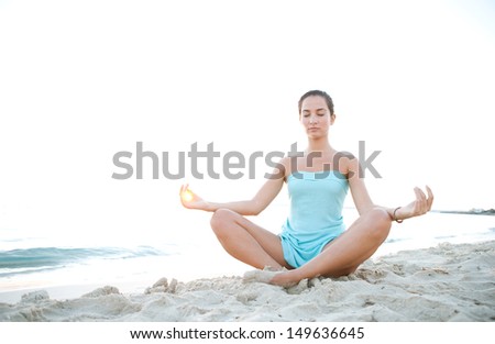 Low angle view of a healthy young woman sitting in a yoga position meditating, with the sun filtering through her fingers in a circle during sunset on a tranquil beach.