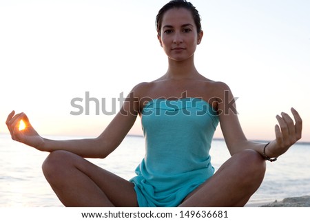 Silhouette view of a healthy young woman sitting in a yoga position meditating, with the sun filtering through her fingers in a circle during sunset on a tranquil beach.
