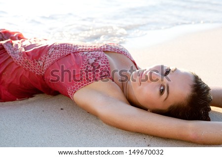 Close up portrait side view of a beautiful woman laying down on a white sand beach with her arms up, bathing in the sea waves dressed in pink at sunset during a summer vacation.