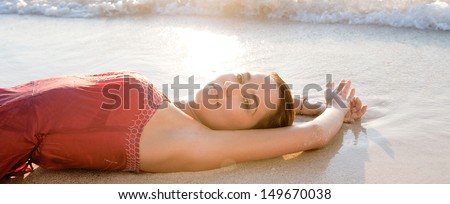 Side view of a young woman laying down on the shore of a white sand beach with her arms up, bathing in the sea waves while dressed in pink at sunset during a summer vacation.