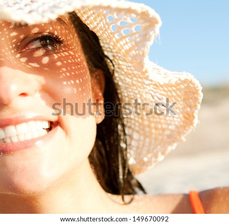 Close up portrait of an attractive smiling woman on vacation on a beach, wearing a straw hat and shading her face with it creating a sun pattern on her skin. Protecting from sun rays.