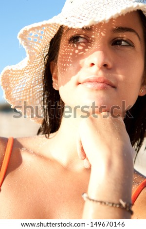 Close up portrait of an attractive woman on vacation on a beach, wearing a straw beam hat and shading her face with it creating a sun pattern on her skin. Protecting from sun rays.