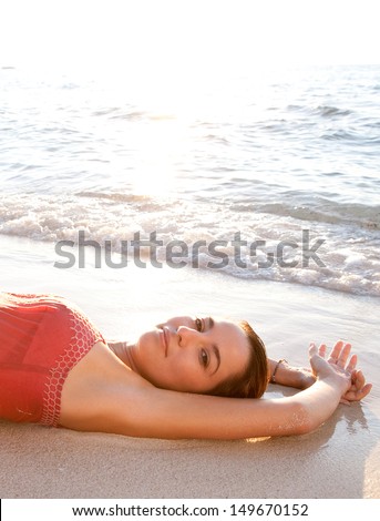 Side view of a beautiful woman laying down on the shore of a white sand beach with her arms up, bathing in the sea waves while dressed in pink at sunset during a summer vacation.