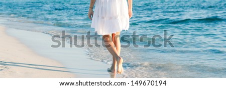 Panoramic rear lower body section view of a woman walking along a white sand beach shore at sunset, taking steps and relaxing with an intense blue sea during a summer vacation.