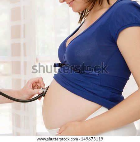 Side view of a pregnant woman middle body section exposing her belly while a gynecologist doctor uses a stethoscope to listen to the baby heart beat at a hospital, indoors.