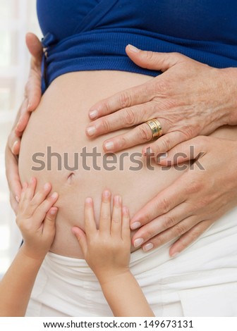 close up detail of a pregnant woman middle body section with a grown belly and the hands of the women in the family generation, grand mother, daughter and grand daughter, home interior.