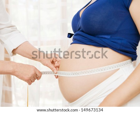 Side view of a pregnant woman middle body section exposing her belly while a gynecologist doctor uses a measuring tape to follow the growth of the baby at a hospital, indoors.