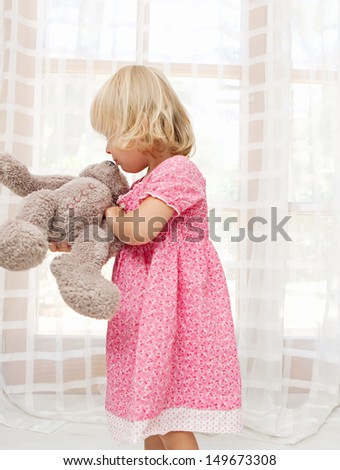 Young girl child playing with her soft teddy bear and kissing it while being at home with white lght curtains and large glass doors during a sunny summer day, interior.