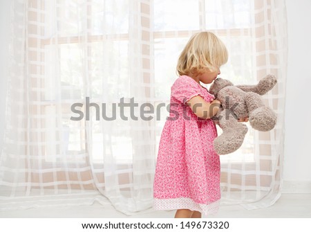 Young Girl Child Playing With Her Soft Teddy Bear And Kissing It While Being At Home With White Light Curtains And Large Glass Doors During A Sunny Summer Day, Interior.