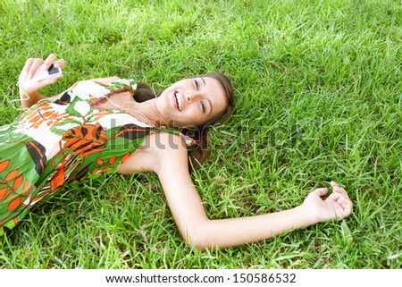 Side portrait view of an attractive young woman laying down on textured green grass listening to music with her mp3 player and smiling joyfully during a summer day outdoors.