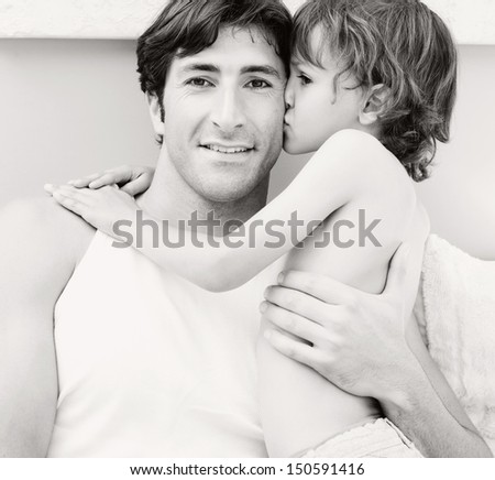Black and white close up portrait of a father and a young son relaxing on a bedroom at home, with the boy kissing the father on the cheek while cuddling him. Home interior.