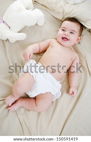 Over head view of a beautiful baby girl laying down on a bed at home with her soft toy friend, joyful and smiling, and wearing a white nappy.