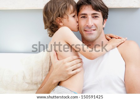 Close up portrait of a father and a young son relaxing on a bedroom at home, with the boy kissing the father on the cheek while cuddling him. Home interior.
