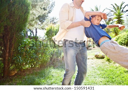 Father playing with his child boy in a home garden during a sunny day, picking him up and flying him turning around and having fun together.