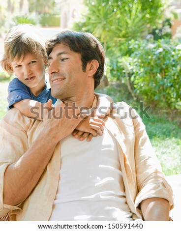Young son hugging his father while relaxing and sitting on a chair by a house garden during a sunny day while on holiday during the summer.