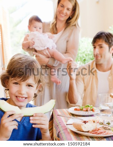 Family of four gathering around a table with food in a home porch garden outdoors with mother, father, baby girl, and a young boy turning to camera and biting a slice of melon.