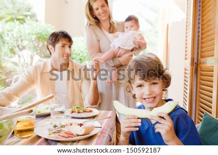 Family of four gathering around a table with food in a home porch garden outdoors with mother, father, baby girl, and a young boy turning to camera and biting a slice of melon.