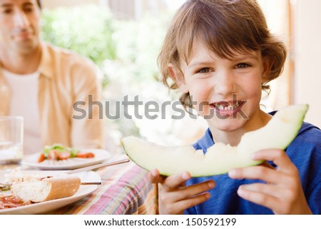 Young kid boy turning to camera and holding a bitten slice of melon while eating outdoors in a home garden porch with his father during a sunny day.