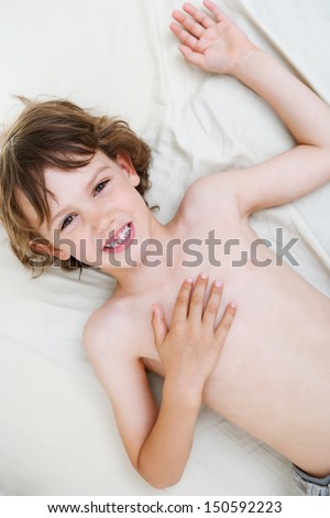 Close up over head portrait of a young boy laying down on a bed at home, smiling and relaxing during a weekend morning with a bare chest, being happy.