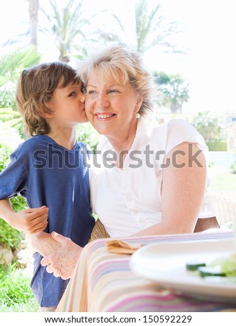Grand child boy kissing grandmother on the cheek while having lunch together in a garden porch at home during a summer sunny day on vacation.