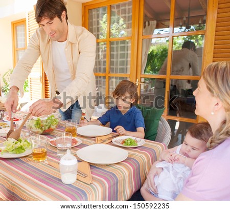 Father of a family of four serving green leaves salad while gathering around a table at lunch, getting ready for a meal while on vacation during a summer day.