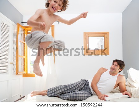 Father Reading At Book In His Bedroom While Young Son Child Is Jumping Up On The Mattress, Having Fun And Being Energetic During A Sunny Weekend Morning At Home.