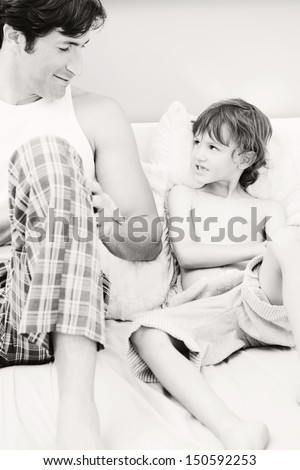 Father and son relaxing together on a bed at home during a weekend morning, bonding and sitting next to each other with the boy copying his father moves, black and white image.