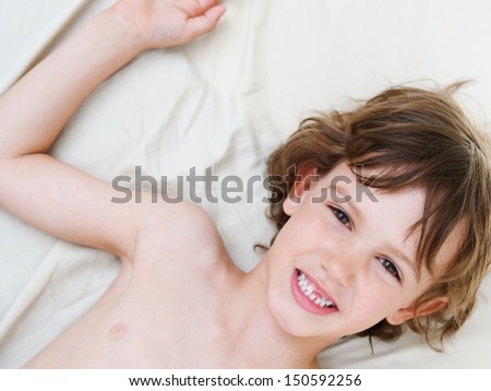 Close up over head portrait of a young boy laying down on a bed at home, smiling and relaxing during a weekend morning with a bare chest, being happy.