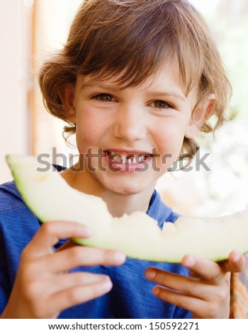 Young boy joyfully holding a bitten slice of melon while eating outdoors in a home garden porch during a sunny day. Close up portrait.