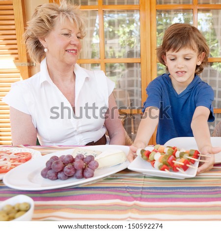Grandmother and grandson sitting together at an outdoors table at home with food and the boy child bringing a plate of vegetable skews, smiling happy.