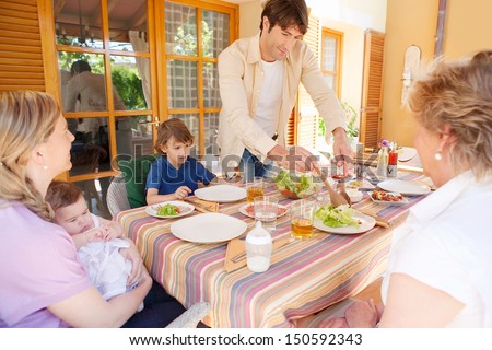 Family of five gathering around a table with food preparing to have lunch, with the father serving green salad during a sunny day outdoors.
