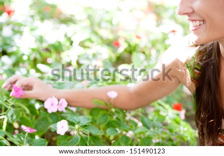 Side view of a young woman in a green park stretching her arm to pick a flower from a bush full of them, being selective and smiling, outdoors.