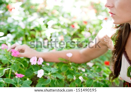 Side view of a young woman in a green park stretching her arm to pick a flower from a bush full of them, being selective, outdoors.