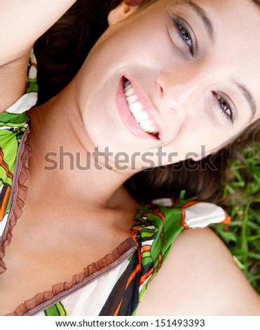 Over head portrait view of a young attractive woman laying down and relaxing on healthy green grass while smiling at the camera during a summer day, outdoors.